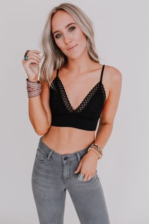 A woman with long blonde hair wears the Audra Waistband Loop Lace Bralette in black with adjustable straps and gray high-waisted jeans. She stands against a plain white background, confidently looking at the camera while wearing bracelets and rings.