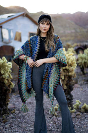 A woman with long hair wears the Everyday Velvet Headband - Black, a colorful knit shawl, black top, gray flared pants, and bracelets. She stands outdoors among desert plants with a rustic building in the background.