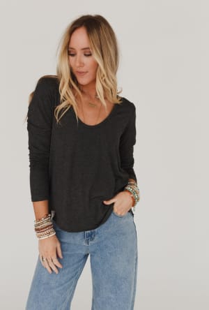 A woman with long blonde hair wears the Ryder Long Sleeve Bralette Tee in Charcoal and light blue jeans, accessorized with colorful bracelets. She stands against a plain white background, looking down and to the side with one hand in her pocket.