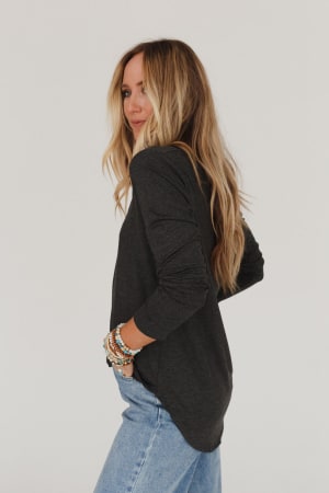 A woman with long blonde hair stands sideways, looking over her shoulder in a Ryder Long Sleeve Bralette Tee - Charcoal and light blue jeans, wearing colorful beaded bracelets, set against a plain white background.