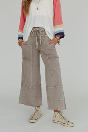 Wearing the Lucky One Wide Leg Pant in taupe, a person with a color-blocked long-sleeve shirt, white high-top sneakers, and bracelets stands against a plain background. Their face is not shown.