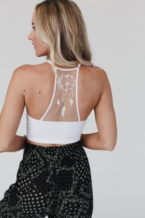 A woman with long blonde hair wears the Peace Dreamcatcher Tattoo Bralette in white, featuring a sheer mesh dreamcatcher design, paired with black patterned pants. She stands with her back to the camera against a plain background.