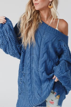 A woman with long blonde hair wears the Golden Hour Bubble Sleeve Sweater Dress in blue, styled off-the-shoulder, over a white tank top and floral pants against a plain light background.