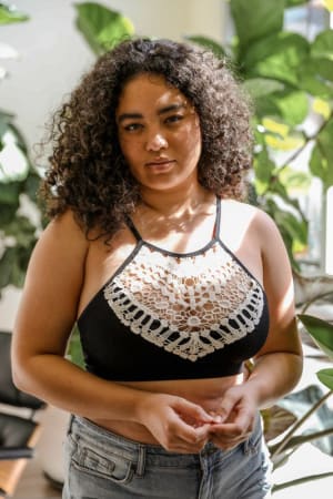 A woman with curly dark hair stands indoors, wearing the Crochet High Neck Bralette in black and light denim jeans, surrounded by green leafy plants and sunlight.