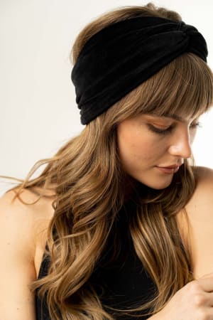 A woman with long, wavy light brown hair wears the Everyday Velvet Headband in Black, featuring subtle criss-cross detailing, paired with a sleeveless black top as she looks down against a neutral background.
