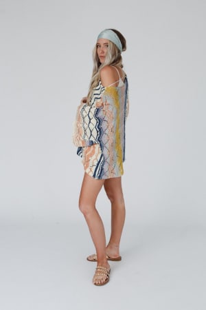 A woman with long, light hair wears the Rainbow Shores Knit Sweater - Multi, paired with a blue headband and pearl-detailed sandals. She stands sideways, looking at the camera against a plain white background.