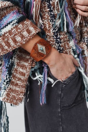Close-up of a person wearing a chunky knit sweater and dark pants, accessorized with the Wynona Diamond Leather Cuff Bracelet in Camel—a wide leather cuff featuring an intricate design and turquoise accent—with one hand in their pocket.