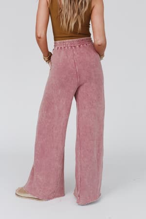 A woman with long, wavy hair stands with her back to the camera against a plain light background, wearing a sleeveless brown top and the Relaxing Robin Wide Leg Pant in New Mauve.