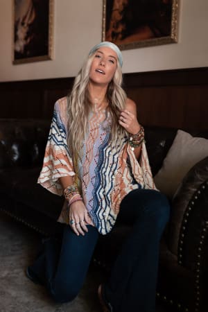 A woman with long blonde hair and a headband kneels by a leather couch in a cozy room, wearing the Rainbow Shores Knit Sweater - Multi with dark flared jeans, accessorized with multiple bracelets and rings. Framed art decorates the space.