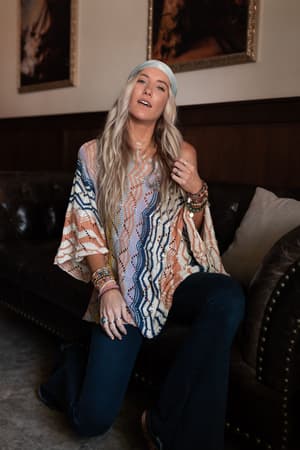 A woman with long blonde hair and a headband kneels by a leather couch in a cozy room, wearing the Rainbow Shores Knit Sweater - Multi with dark flared jeans, accessorized with multiple bracelets and rings. Framed art decorates the space.