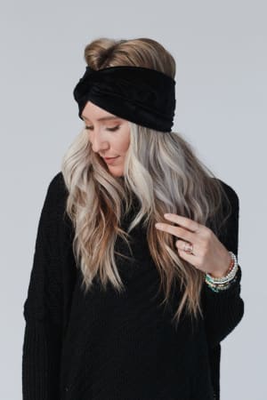 A woman with long, wavy blonde hair wears the Everyday Velvet Headband - Black with criss-cross detailing and a black sweater. She looks down, touching her hair and displaying several bracelets and rings against a light background.