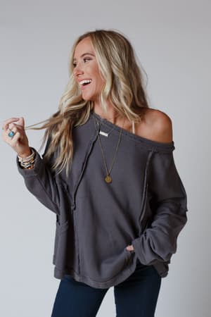 A woman with long blonde hair smiles and looks to the side, wearing the Bailey Oversized Raw Edge Top in charcoal with blue jeans and layered jewelry, softly holding a lock of her hair against a plain background.
