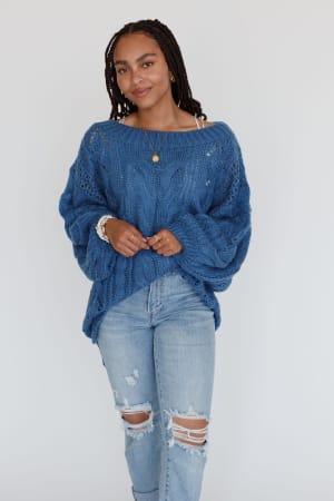A woman with braided hair wears the Golden Hour Bubble Sleeve Sweater Dress in blue, paired with layered necklaces and bracelets. She stands against a plain white background, smiling slightly.
