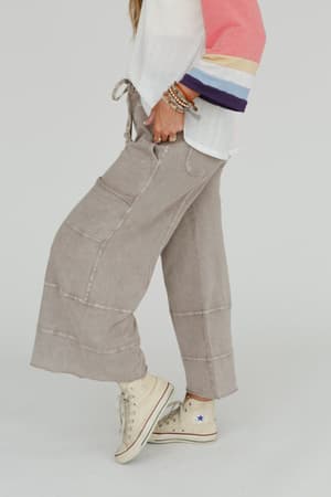 Wearing the Lucky One Wide Leg Pant in taupe, a white and pink color-blocked long-sleeve top, stacked beaded bracelets, and off-white high-top Converse sneakers, a person stands against a plain background.