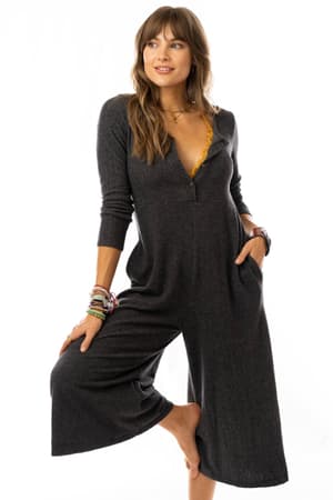 A barefoot woman with long brown hair, layered necklaces, and colorful bracelets wears the Simply Comfort Jumpsuit - Dark Gray with long sleeves, a deep V-neck, and pockets. She lifts one leg and smiles slightly.