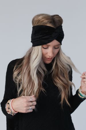A woman with long, wavy blonde hair wears the Everyday Velvet Headband - Black, featuring criss-cross detailing, and a textured sweater. She looks down, displaying beaded bracelets and rings against a plain background.