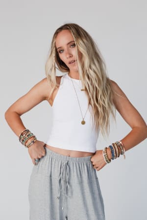 A woman with long, wavy blonde hair wears the High Neck Racerback Brami in white with gray drawstring pants, layered bracelets, and a necklace. She stands against a plain background with her hands on her hips.