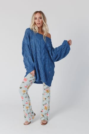 A woman with long blonde hair wears the Golden Hour Bubble Sleeve Sweater Dress in blue, styled off one shoulder, paired with floral light blue bell-bottom pants and sandals, standing against a plain white background.
