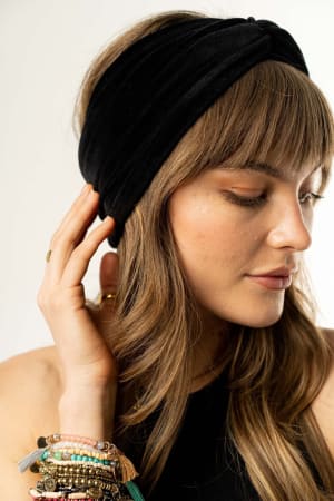 A woman with long, wavy hair wears the Everyday Velvet Headband in Black, featuring criss-cross detailing. She gently touches the accessory and looks down softly, with several beaded bracelets on her left wrist against a plain background.