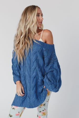 A woman with long blonde hair wears the Golden Hour Bubble Sleeve Sweater Dress in blue, styled over a white top and floral leggings. She looks to the side against a plain, light background.