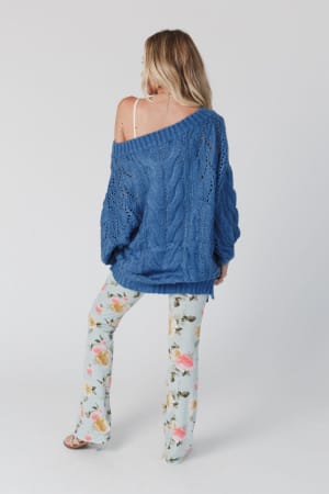 A woman with long blonde hair poses relaxed against a plain background, wearing the Golden Hour Bubble Sleeve Sweater Dress in blue off one shoulder, exposing a white strap, paired with light blue floral pants.