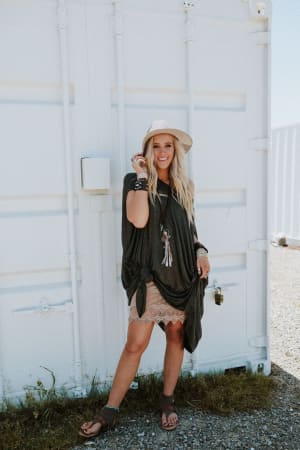 A woman stands outdoors by a white metal wall, smiling. She wears a wide-brim hat, a dark top with tassel necklaces, sandals, bracelets, and the Layering Love Lace Slip - Mocha under her lace-trimmed skirt, holding her hat with one hand.