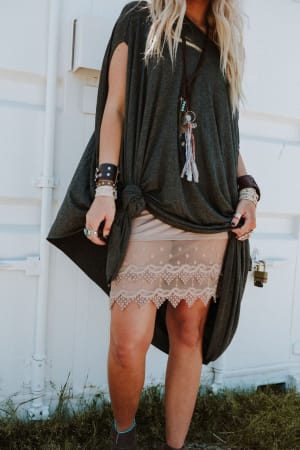 A person wears the Layering Love Lace Slip - Mocha under a loose dark gray tunic, styled with layered necklaces, bracelets, and rings, posing outside against a white wall.
