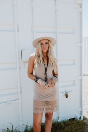 A woman with long blonde hair wears the Layering Love Lace Slip in mocha and a wide-brimmed hat. She stands outdoors before a white metal container on a sunny day, gazing at the camera and accessorized with multiple bracelets.