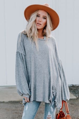 A woman with long blonde hair wears the Charlie Cozy Top in gray—an oversized sweater with ruffled edges and puff sleeves—paired with ripped light blue jeans, a tan wide-brim hat, and a brown handbag, standing outdoors by a white wall.
