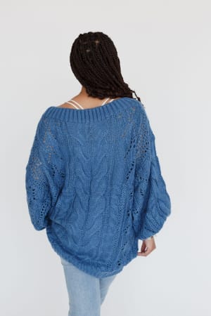 A woman with braided hair, seen from behind, wears the Golden Hour Bubble Sleeve Sweater Dress in blue over a white top and light blue jeans against a plain white background.