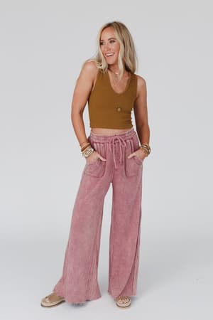 A woman smiles with her hands in the drawstring waist of the Relaxing Robin Wide Leg Pant in New Mauve. She wears a brown sleeveless top, layered necklaces, several bracelets, and sandals against a plain light background.