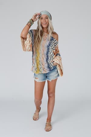 A smiling woman with long blonde hair models the Rainbow Shores Knit Sweater - Multi, featuring a colorful boho design, slouchy loose fit, and flare sleeves, paired with ripped denim shorts and accessories against a plain white background.