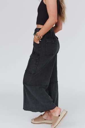 A woman stands sideways against a plain light background wearing the Game On Cargo Wide Leg Pant in black with a sleeveless crop top, tan espadrille sandals, and beaded bracelets.
