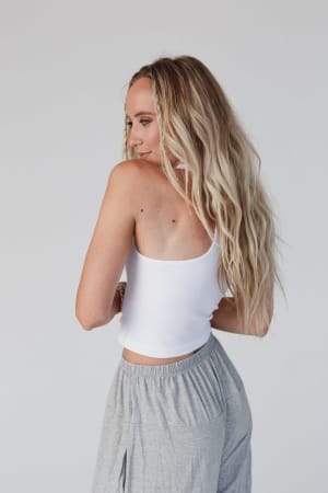 A woman with long, wavy blonde hair models the white High Neck Racerback Brami, paired with gray pants, as she stands sideways and looks over her shoulder against a plain light background.