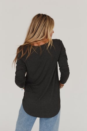 A woman with long blonde hair stands with her back to the camera, wearing the Ryder Long Sleeve Bralette Tee in charcoal and light blue jeans against a plain white background.