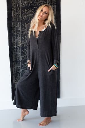 A woman with long blonde hair stands barefoot on a white floor, wearing the Simply Comfort Jumpsuit in dark gray with an oversized fit. She has her hands in her pockets and colorful bracelets, against a backdrop of a dark patterned tapestry.