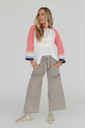 A woman stands confidently in the Lucky One Wide Leg Pant - Taupe, paired with a loose-fitting white and pink long-sleeve shirt, beige sneakers, layered bracelets, a gold necklace, and long blonde hair against a plain background.