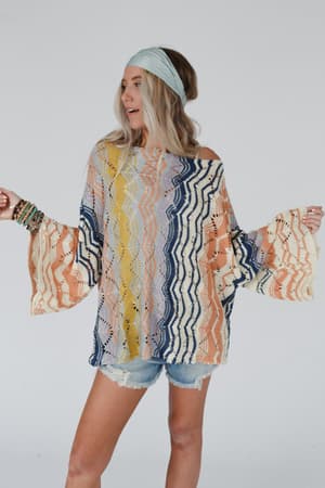 A woman in a light blue headband and the Rainbow Shores Knit Sweater - Multi with wavy patterns and distressed denim shorts stands against a plain background, holding her arms out to the sides.