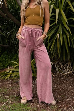 A woman stands outdoors in front of green plants, wearing a mustard crop top, sandals, layered necklaces, bracelets, and the Relaxing Robin Wide Leg Pant in New Mauve with a drawstring waist, her hands casually in her pockets.
