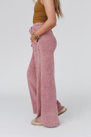 A woman stands sideways wearing the Relaxing Robin Wide Leg Pant in New Mauve with a drawstring waist, loose fit, and pockets. She pairs it with a sleeveless brown ribbed top and sandals; her hair is down against a plain white background.