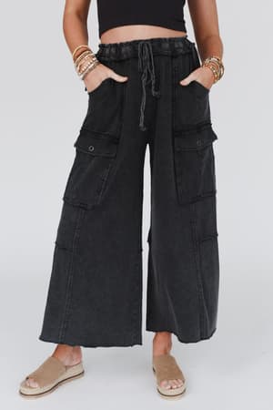 Person wearing Game On Cargo Wide Leg Pant - Black with a black crop top, bracelets, and beige open-toe espadrille sandals, standing against a plain light background.