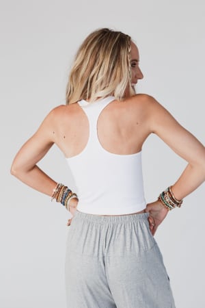 A woman with long blonde hair wears the High Neck Racerback Brami in white, paired with gray pants and beaded bracelets. She stands hands on hips, facing away against a plain light background.