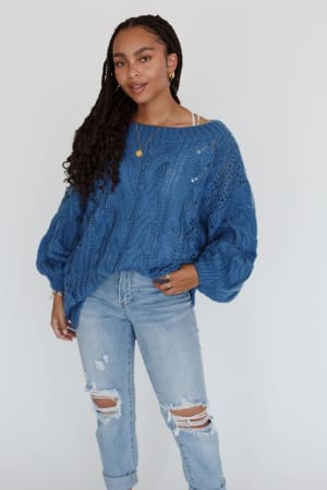 A woman with long braided hair confidently poses in a Golden Hour Bubble Sleeve Sweater Dress - Blue, accessorized with a gold necklace and ring, against a plain white background.