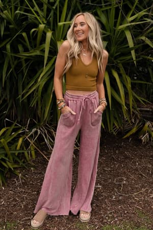 A woman with long blonde hair, wearing a brown tank top, sandals, and the Relaxing Robin Wide Leg Pant in New Mauve with a drawstring waist, stands outdoors in front of lush green plants, smiling with her hands in her pockets.