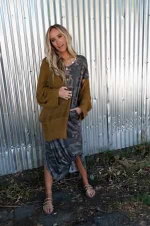 A woman with long blonde hair poses casually in front of a corrugated metal wall, wearing a camo-print dress, layered necklaces, studded sandals, and the Brenden Knit Netted Cardigan in brown.