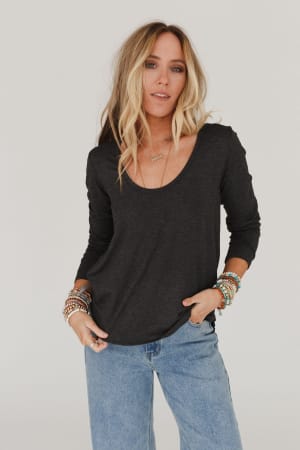 A woman with long, wavy blonde hair wears the Ryder Long Sleeve Bralette Tee in Charcoal and light blue jeans, accessorized with bracelets and a necklace, standing against a plain light background.