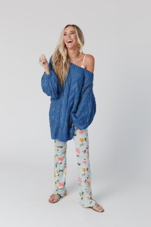 A woman with long blond hair smiles while wearing the Golden Hour Bubble Sleeve Sweater Dress in blue and sandals, standing against a plain white background.