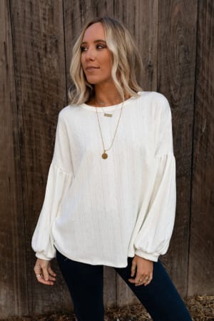 A woman with blonde hair wears The Nest Sweet Heart Balloon Sleeve Knit Sweater in Cream with dark jeans and layered gold necklaces as she stands before a rustic wooden wall, gazing to the side.