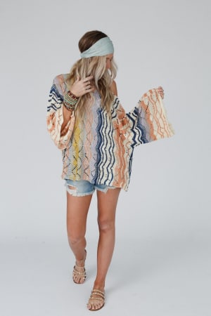 A woman wears the Rainbow Shores Knit Sweater - Multi with its colorful boho style, slouchy fit, and flare sleeves. She pairs it with denim shorts, sandals, a blue headband, and bracelets while standing against a white background.