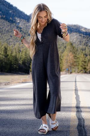 A woman with long, wavy blonde hair smiles while walking on a road in the Simply Comfort Jumpsuit - Dark Gray, paired with white sandals. Mountains and trees create a scenic backdrop under a clear sky.